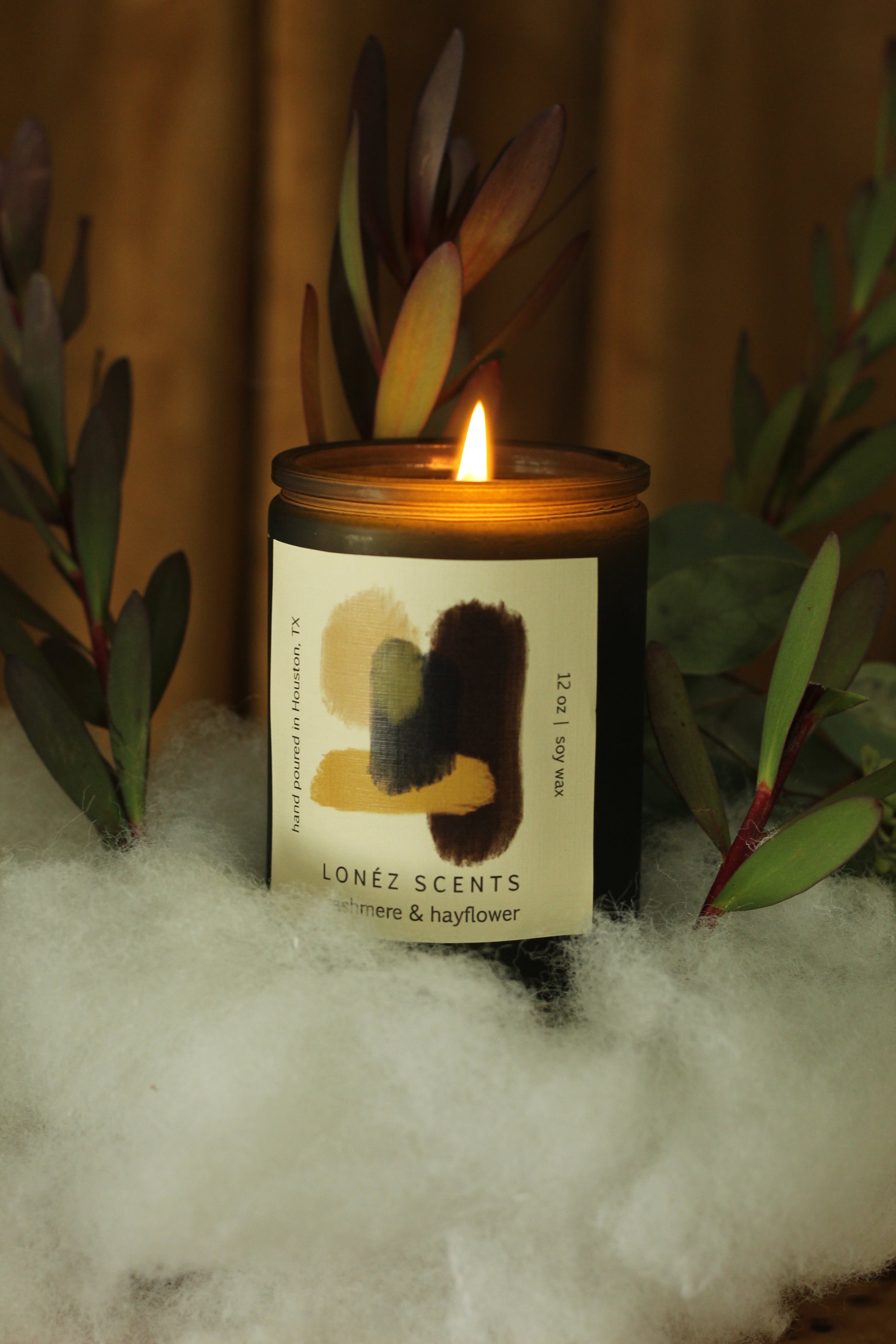 Cashmere & hayflower scented candle.  The candle is in a deep green votive. The candle is propped on a small installation made of poly-fill with the intention oc depicitng a cloud. There is also greenery sticking out of the cloud. The backround is a rust colored curtain.